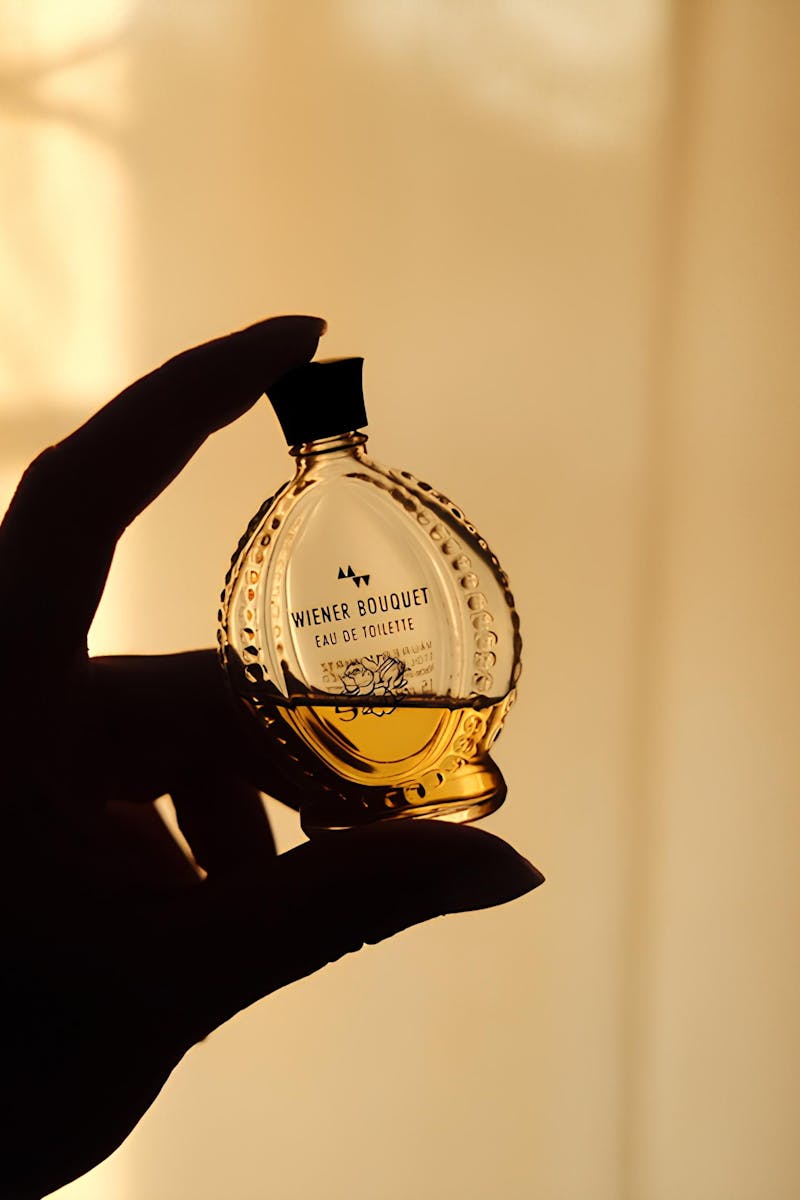 A silhouette of an elegant perfume bottle held by a hand against a warm, dimly lit background.